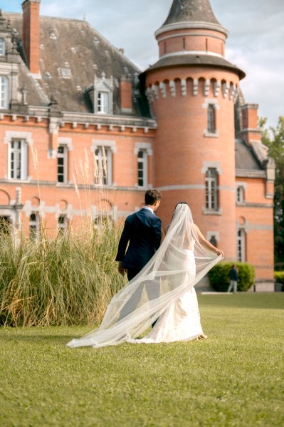 Bride has hand in the air walking hand in hand with groom on grass veil is blowing in the wind